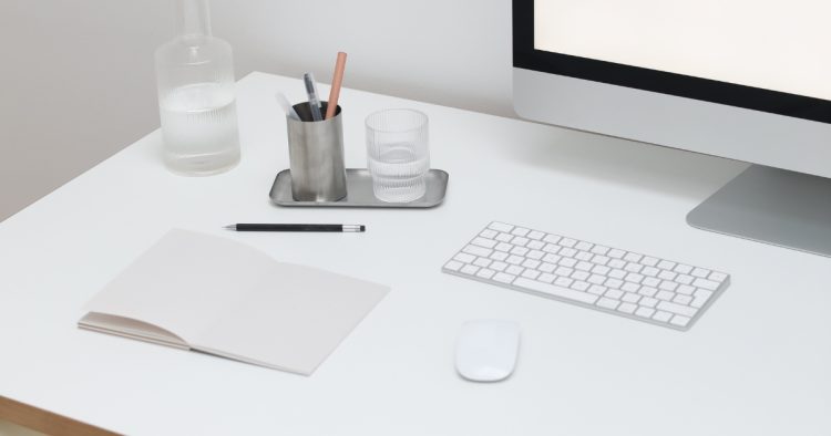 A clean, minimalist white workspace featuring a keyboard, computer monitor, pen, and open notebook.