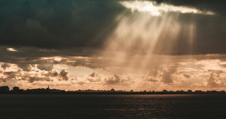 Sunlight breaking through dark storm clouds over water, representing clarity and overcoming imposter syndrome.