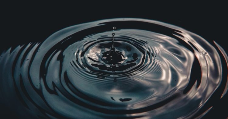 A single water droplet creating expanding concentric ripples on dark water, symbolizing the widespread impact of ethical leadership.