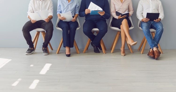 Five job candidates holding CVs sitting in a row in an office waiting room, representing the competitive interview and selection process.