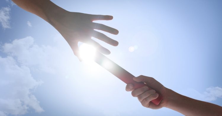 Two hands passing a relay baton against a sunlit blue sky, representing seamless recruitment handoffs and effective candidate evaluation.