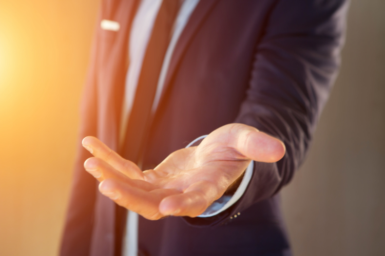 A businessman in a suit holding out an open palm illuminated by bright sunlight, representing opportunity and financial growth in a self-employed sales career.