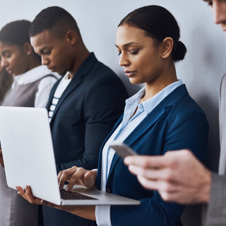 A diverse group of professional job applicants standing in line reviewing laptops and phones ahead of an interview.