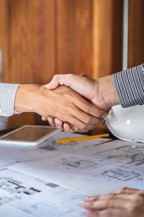 Two business professionals shaking hands over plans at a desk, representing mutual agreement on a sales career ladder and commission plan.