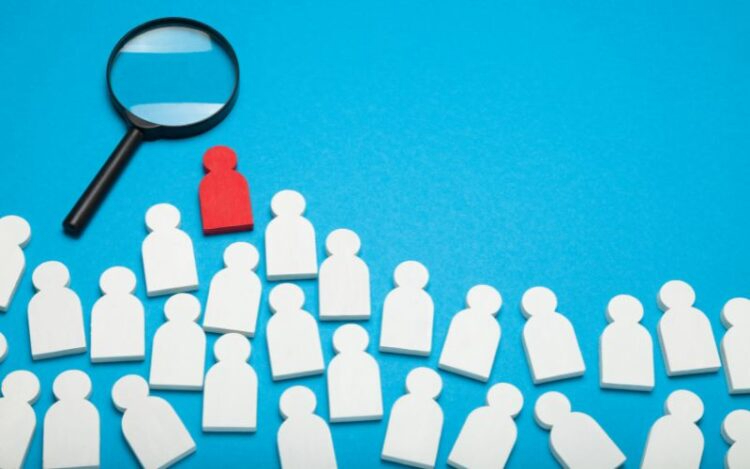 A magnifying glass focusing on a single red figure among white wooden figures on a blue background, illustrating how to attract self-employed sales talent.