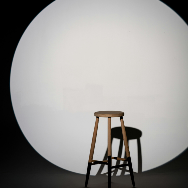 A single wooden stool standing illuminated under a circular spotlight casting a dark shadow on a backdrop.
