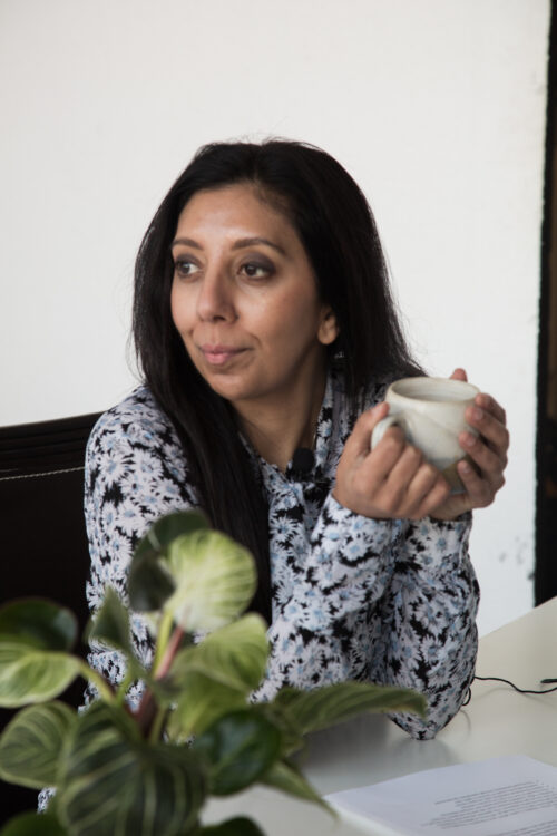 Leena Parmar drinking from a tea cup sat at a desk behind a plant