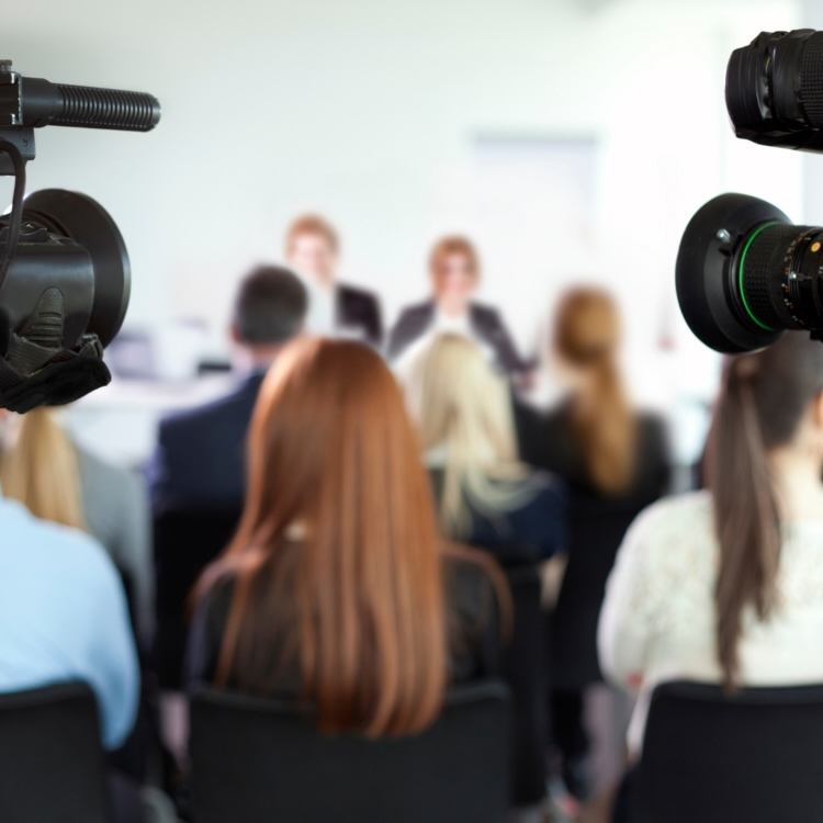 Professional video cameras recording a media press conference with speakers presenting to an audience.