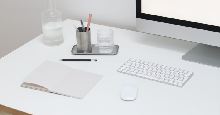 A clean, minimalist white workspace featuring a keyboard, computer monitor, pen, and open notebook.