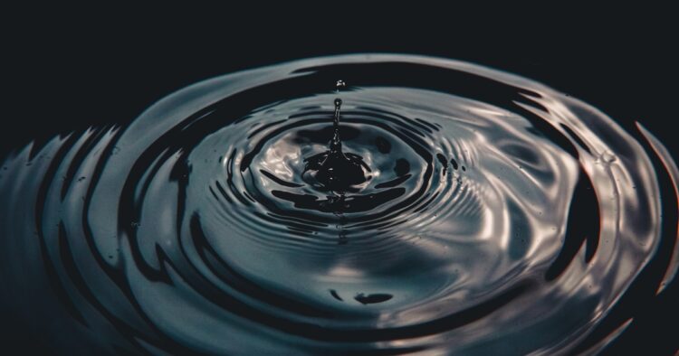 A single water droplet creating expanding concentric ripples on dark water, symbolizing the widespread impact of ethical leadership.