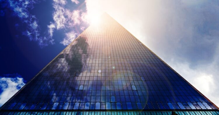 Low-angle view looking up a modern glass skyscraper reflecting clouds and sunlight, representing UK B2C sales market growth and recruitment trends.