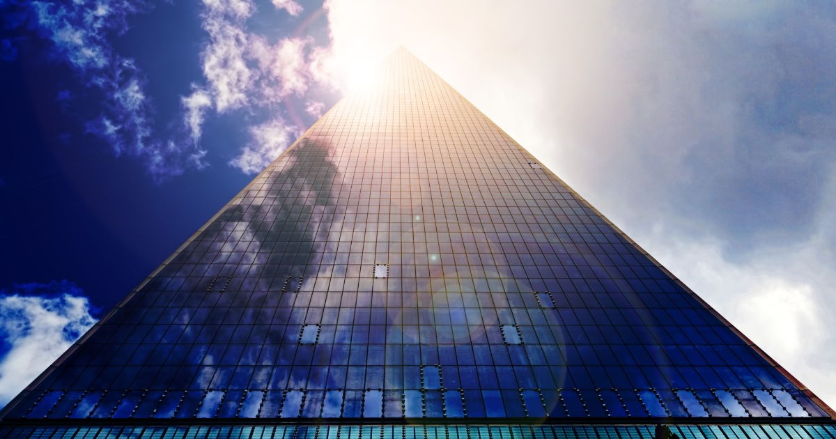 Low-angle view looking up a modern glass skyscraper reflecting clouds and sunlight, representing UK B2C sales market growth and recruitment trends.